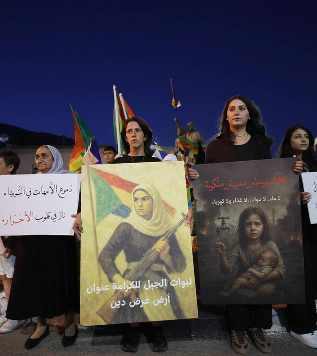 Mehrere Menschen halten Banner und Plakate bei einer Demonstration im Dunkeln.
