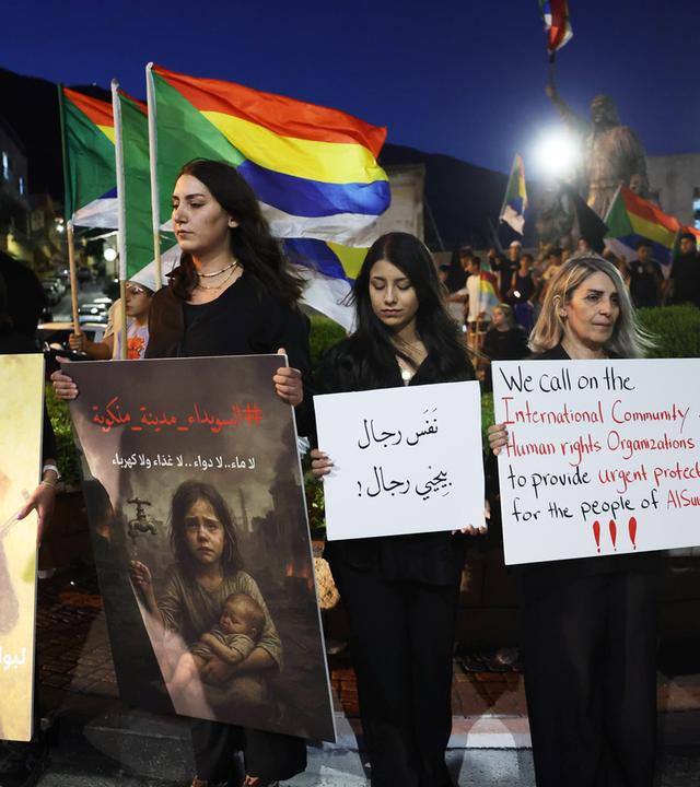 Mehrere Menschen halten Banner und Plakate bei einer Demonstration im Dunkeln.
