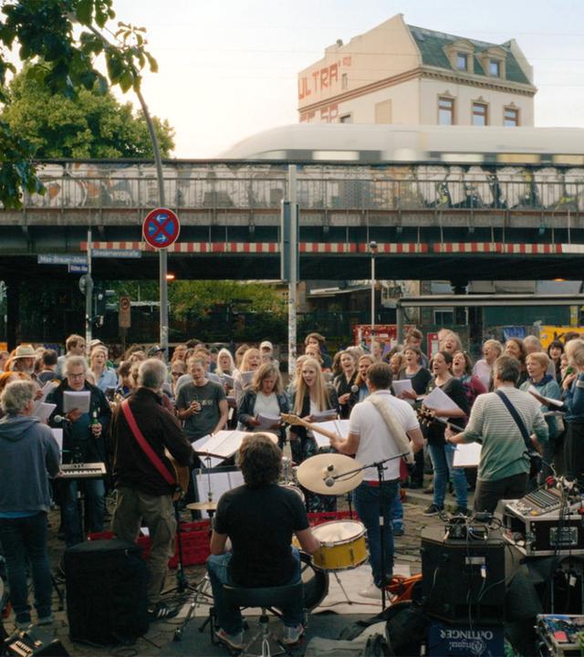 Im Vordergrund stehen eine Band und viele Menschen, die zusammen singen und Musik machen. Im Hintergrund ist eine Brücke zu sehen, über die ein Zug fährt.