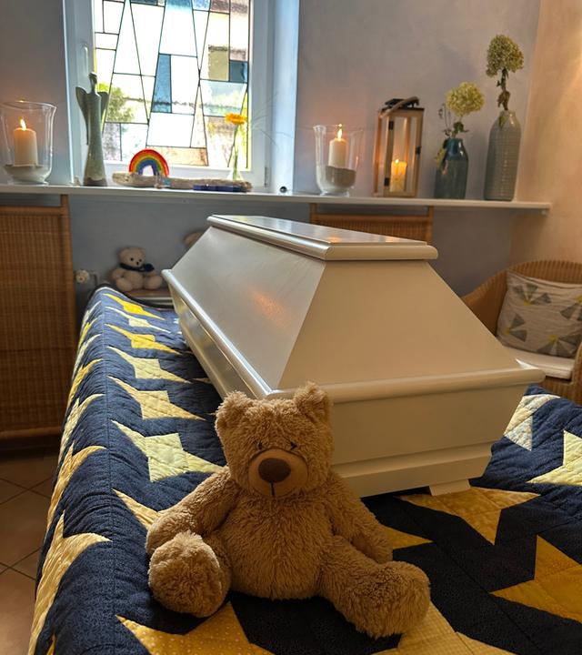 Heller Kindersarg auf einer Sternendecke, davor ein Teddy, im Hintergrund ein Buntglasfenster und brennende Windlichter.