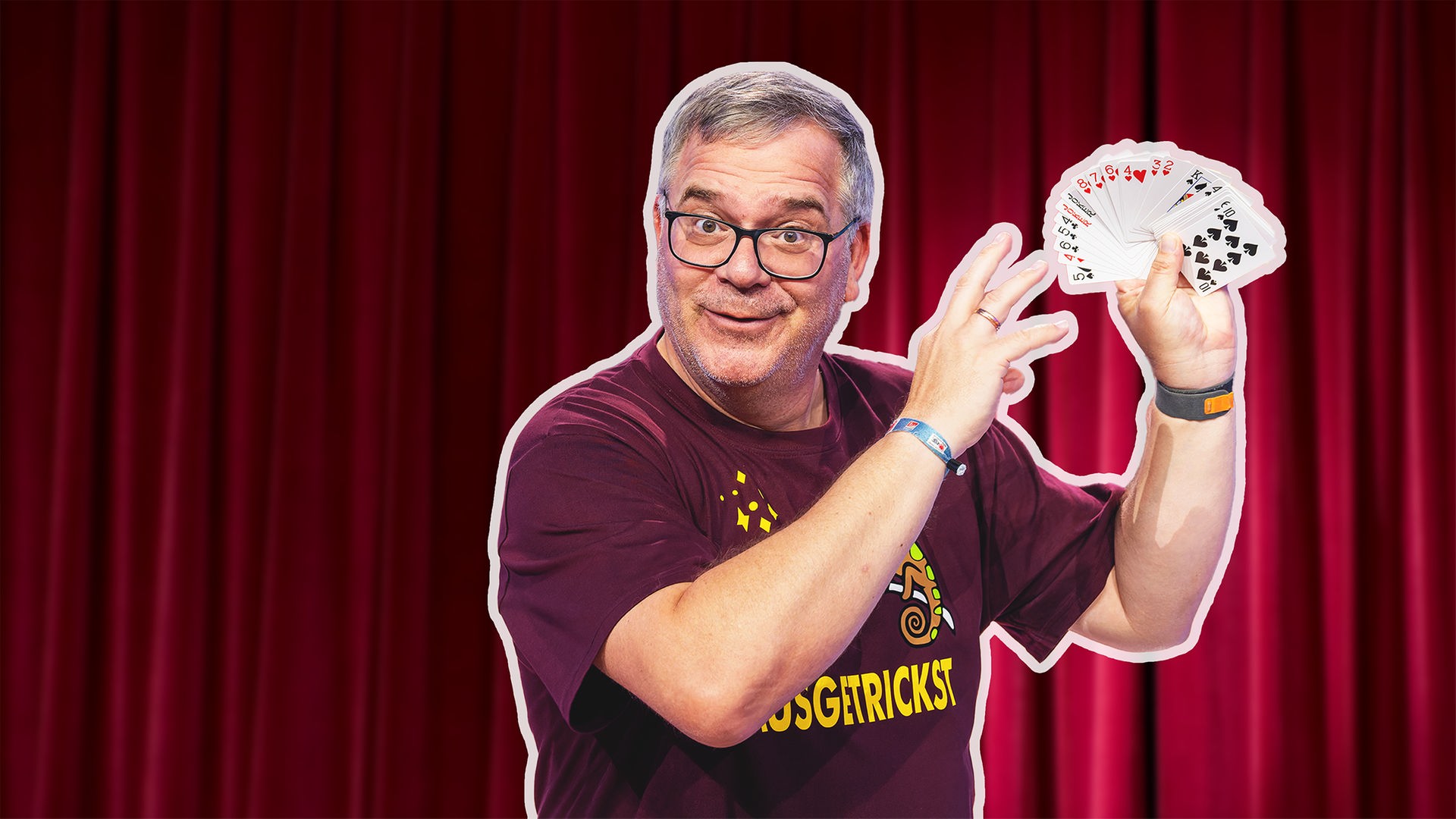 Moderator Elton steht vor einem roten Bühnenvorhang und hält ein aufgefächertes Kartenspiel in der Hand. Er lächelt in die Kamera und zeigt mit der anderen Hand auf die Karten, als würde er einen Trick präsentieren.
