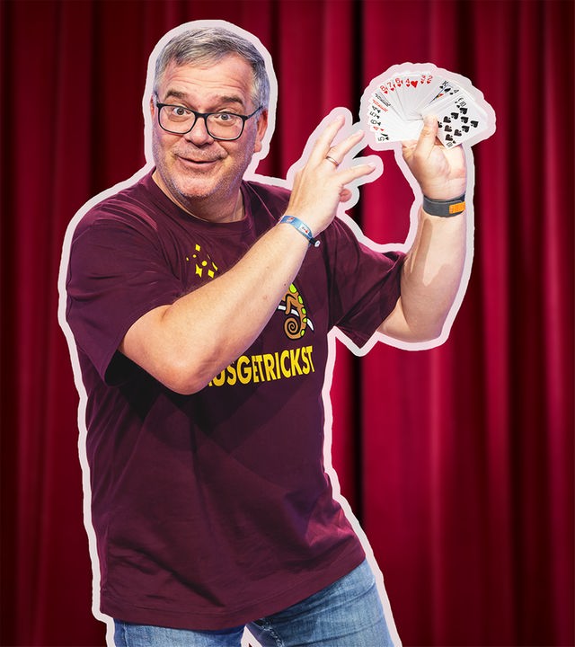 Moderator Elton steht vor einem roten Bühnenvorhang und hält ein aufgefächertes Kartenspiel in der Hand. Er lächelt in die Kamera und zeigt mit der anderen Hand auf die Karten, als würde er einen Trick präsentieren.