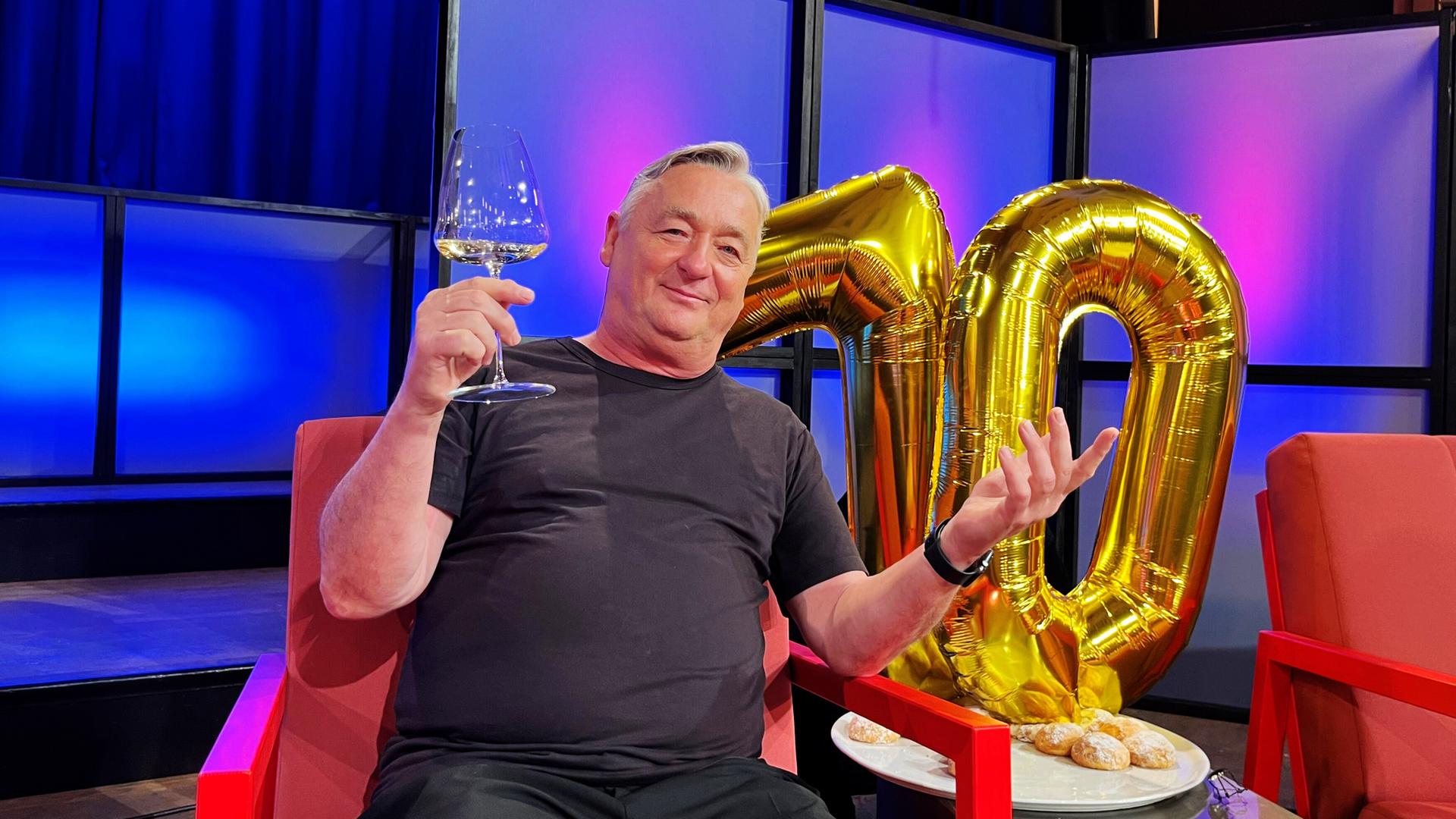 Ein Mann sitzt mit Weinglas in der Hand vor einer goldenen Zahl.