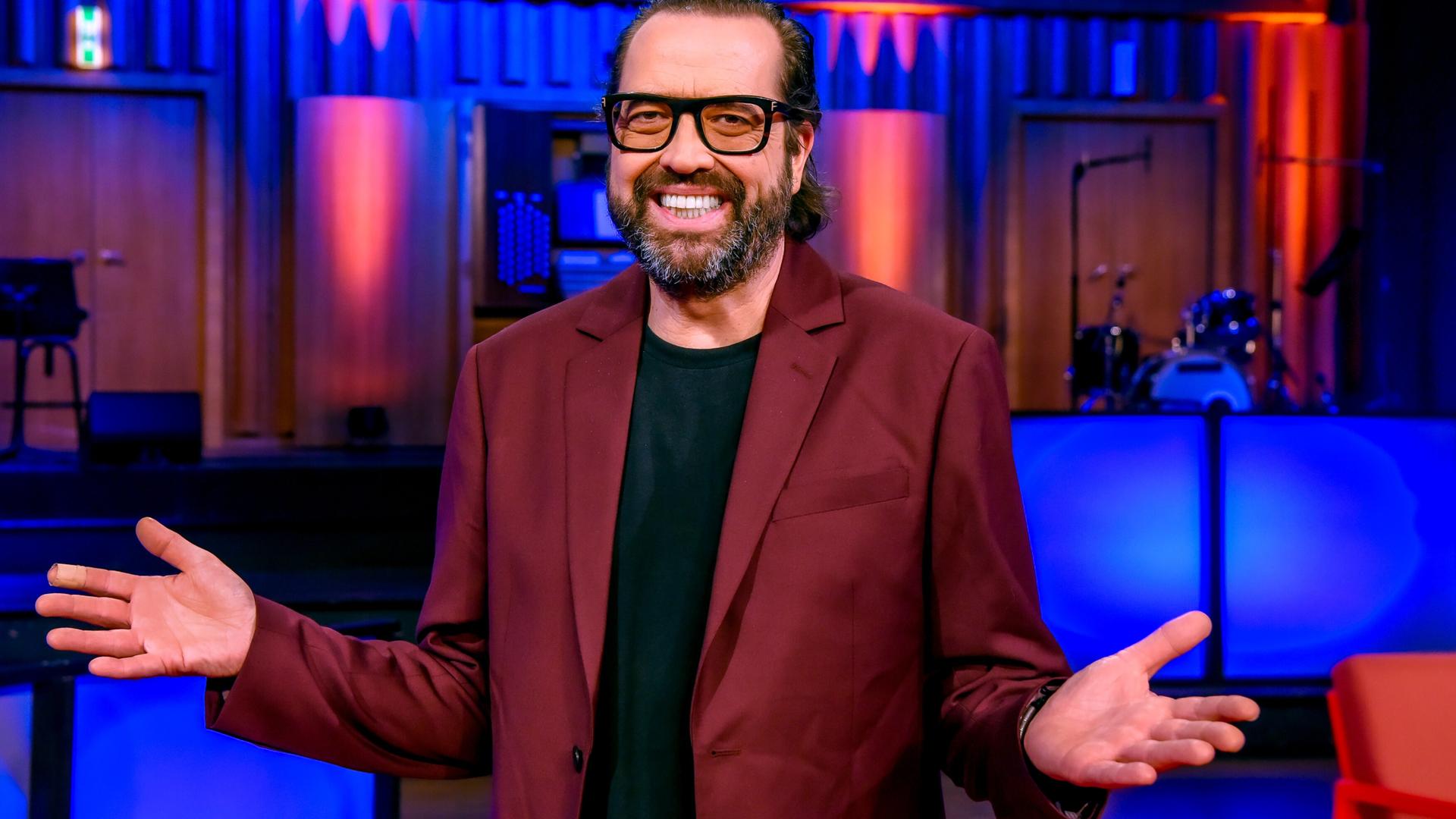 Das Bild zeigt einen Mann, der in einem Raum mit einer Bühne und farbigen Lichtern steht. Er trägt einen rotbraunen Blazer über einem schwarzen T-Shirt. Der Mann hat einen kurzen, gepflegten Bart und trägt eine Brille. Er macht eine offene Handbewegung mit seinen Armen, was Freundlichkeit und Offenheit ausdrückt. Im Hintergrund sind unscharf einige Instrumente und Bühnenausstattungen zu erkennen, die auf eine Unterhaltungssituation hindeuten. Die Atmosphäre wirkt lebhaft und einladend.