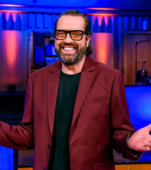 Das Bild zeigt einen Mann, der in einem Raum mit einer Bühne und farbigen Lichtern steht. Er trägt einen rotbraunen Blazer über einem schwarzen T-Shirt. Der Mann hat einen kurzen, gepflegten Bart und trägt eine Brille. Er macht eine offene Handbewegung mit seinen Armen, was Freundlichkeit und Offenheit ausdrückt. Im Hintergrund sind unscharf einige Instrumente und Bühnenausstattungen zu erkennen, die auf eine Unterhaltungssituation hindeuten. Die Atmosphäre wirkt lebhaft und einladend.