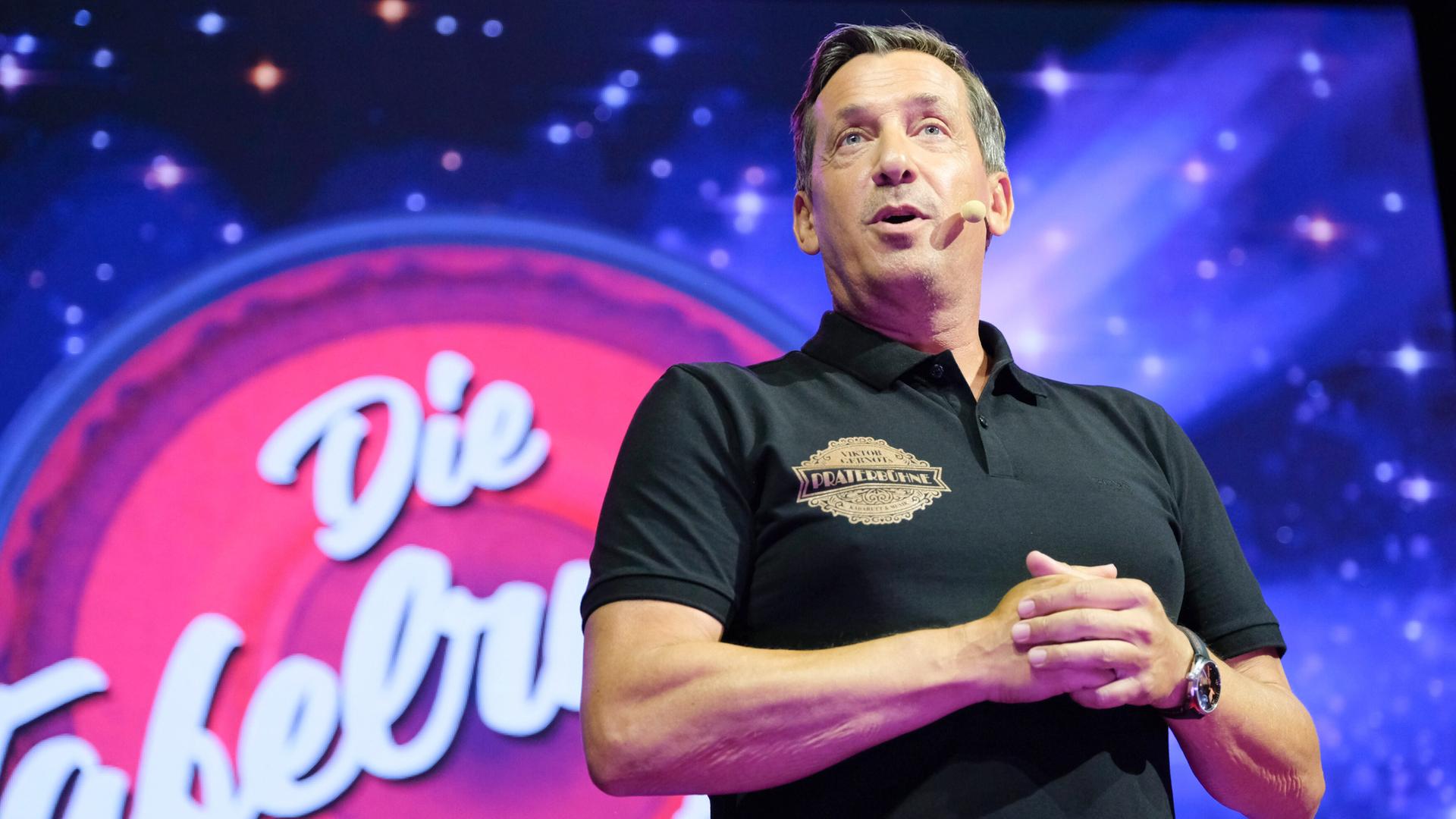 Das Bild zeigt einen Mann, der auf einer Bühne steht und spricht. Er trägt ein schwarzes Poloshirt mit einem Aufdruck, der das Wort "Praterbühne" zeigt. Im Hintergrund ist eine große, farbenfrohe LED-Leinwand, die mit Sternen und einem rot-rosa Farbton gestaltet ist. Auf der Leinwand steht der Schriftzug "Die Tafelrunde". Die Beleuchtung ist hell und erzeugt eine festliche Atmosphäre. Der Mann hat kurzes, leicht graues Haar und schaut ins Publikum, während er seine Hände in einer gestikulierenden Position hält. Es handelt sich um eine Live-Performancesituation.