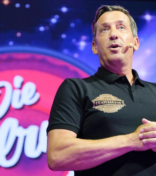 Das Bild zeigt einen Mann, der auf einer Bühne steht und spricht. Er trägt ein schwarzes Poloshirt mit einem Aufdruck, der das Wort "Praterbühne" zeigt. Im Hintergrund ist eine große, farbenfrohe LED-Leinwand, die mit Sternen und einem rot-rosa Farbton gestaltet ist. Auf der Leinwand steht der Schriftzug "Die Tafelrunde". Die Beleuchtung ist hell und erzeugt eine festliche Atmosphäre. Der Mann hat kurzes, leicht graues Haar und schaut ins Publikum, während er seine Hände in einer gestikulierenden Position hält. Es handelt sich um eine Live-Performancesituation.