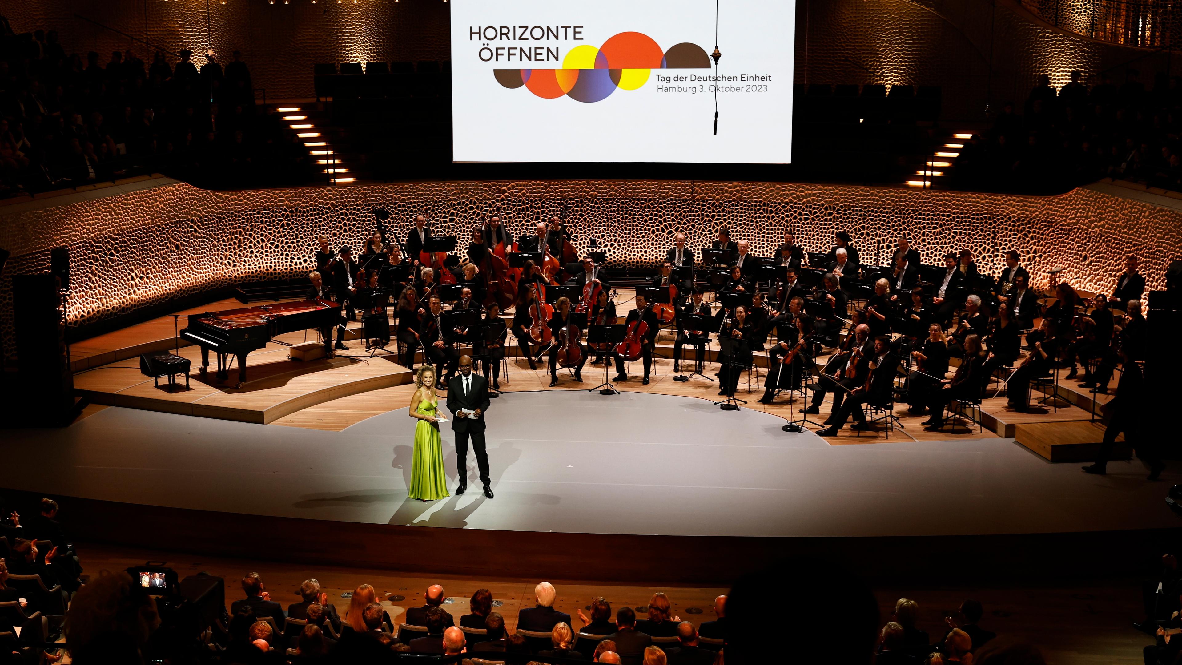 ei Blick von oben auf die Bühne beim Festakt zum Tag der deutschen Einheit 2023, vorne die Moderatoren, hinten ein Orchester.
