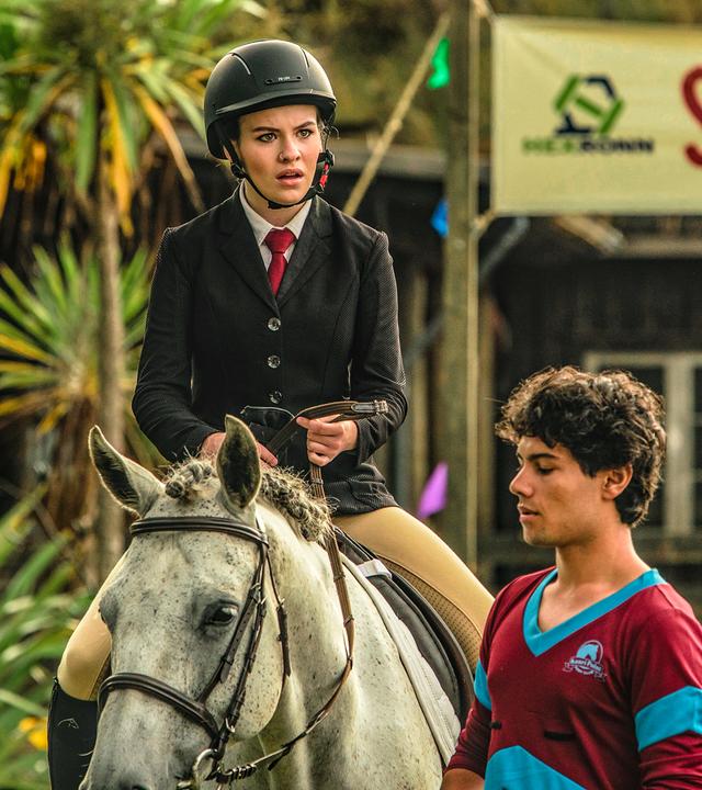 Issie sitzt auf einem Pferd, einem weißen Schimmel. Sie ist kurz davor, beim Reitfest anzutreten und hat einen entrüsteten Blick aufgesetzt. Neben ihr und dem Pferd steht Dan.