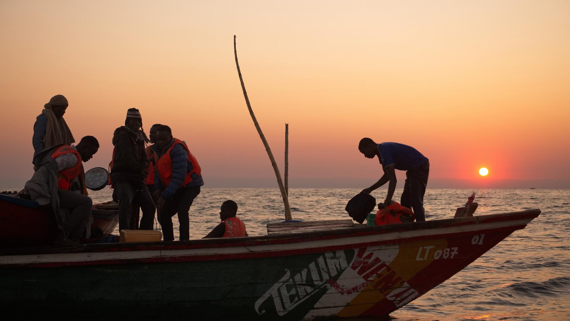 Das Bild zeigt eine Gruppe von sieben Personen auf einem Boot im Tanganjikasee, während die Sonne am Horizont aufgeht oder untergeht und einen warmen farblichen Verlauf im Himmel erzeugt, von Orange bis Violett. Die Personen tragen orangefarbene Schwimmwesten und scheinen mit Fischereiausrüstung zu hantieren. Einzelne Menschen sind dabei, Taschen und Utensilien zu bewegen, während eine Person am hinteren Ende des Bootes steht. Das Boot ist farbig gestaltet, mit einem deutlich sichtbaren Schriftzug „TEKUM WENZA“ an der Seite. Der Wasserstand des Tanganjikasees ist relativ ruhig und spiegelnd, was die Umgebung reflektiert. Im Hintergrund sind sanfte Wellen zu sehen, und der Horizont ist klar, ohne sichtbar nennenswerte Landformationen. Die Stimmung des Bildes vermittelt ein Gefühl der Ruhe und der Verbundenheit mit der Natur.