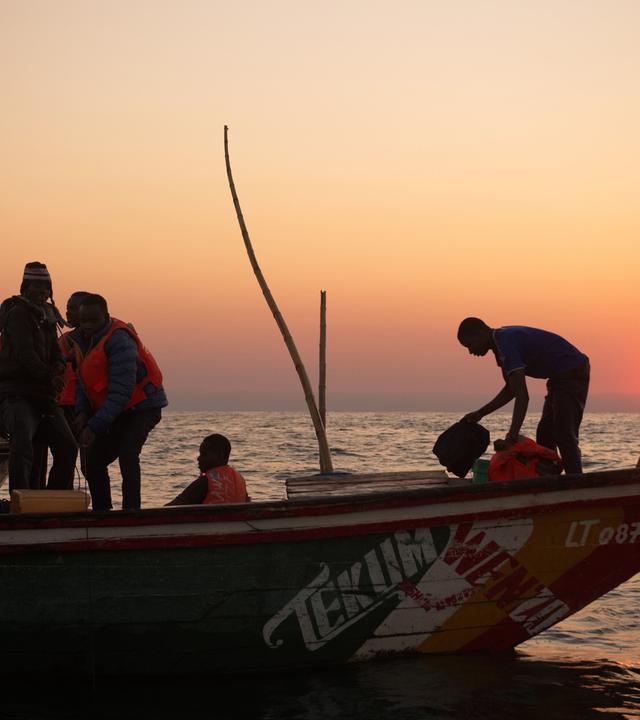 Das Bild zeigt eine Gruppe von sieben Personen auf einem Boot im Tanganjikasee, während die Sonne am Horizont aufgeht oder untergeht und einen warmen farblichen Verlauf im Himmel erzeugt, von Orange bis Violett. Die Personen tragen orangefarbene Schwimmwesten und scheinen mit Fischereiausrüstung zu hantieren. Einzelne Menschen sind dabei, Taschen und Utensilien zu bewegen, während eine Person am hinteren Ende des Bootes steht. Das Boot ist farbig gestaltet, mit einem deutlich sichtbaren Schriftzug „TEKUM WENZA“ an der Seite. Der Wasserstand des Tanganjikasees ist relativ ruhig und spiegelnd, was die Umgebung reflektiert. Im Hintergrund sind sanfte Wellen zu sehen, und der Horizont ist klar, ohne sichtbar nennenswerte Landformationen. Die Stimmung des Bildes vermittelt ein Gefühl der Ruhe und der Verbundenheit mit der Natur.