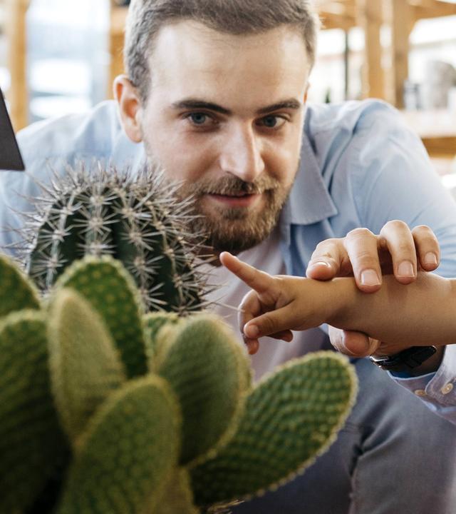 Ein Kind testet in einem Gartencenter einen Kaktus mit dem Finger. Links und rechts daneben sind Vater und Mutter. 