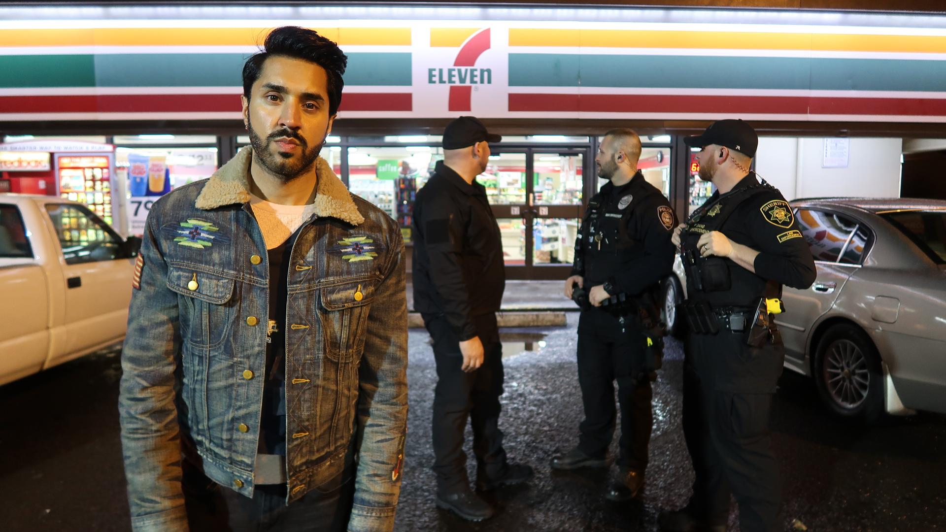 "Tatmotiv Rassismus - Der Tod des Larnell Bruce": Der Journalist Mobeen Azhar steht auf dem Parkplatz vor einem 7-Eleven-Supermarkt. Im Hintergrund stehen drei Polizisten.