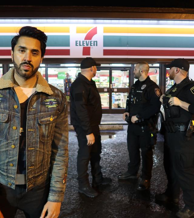 "Tatmotiv Rassismus - Der Tod des Larnell Bruce": Der Journalist Mobeen Azhar steht auf dem Parkplatz vor einem 7-Eleven-Supermarkt. Im Hintergrund stehen drei Polizisten.