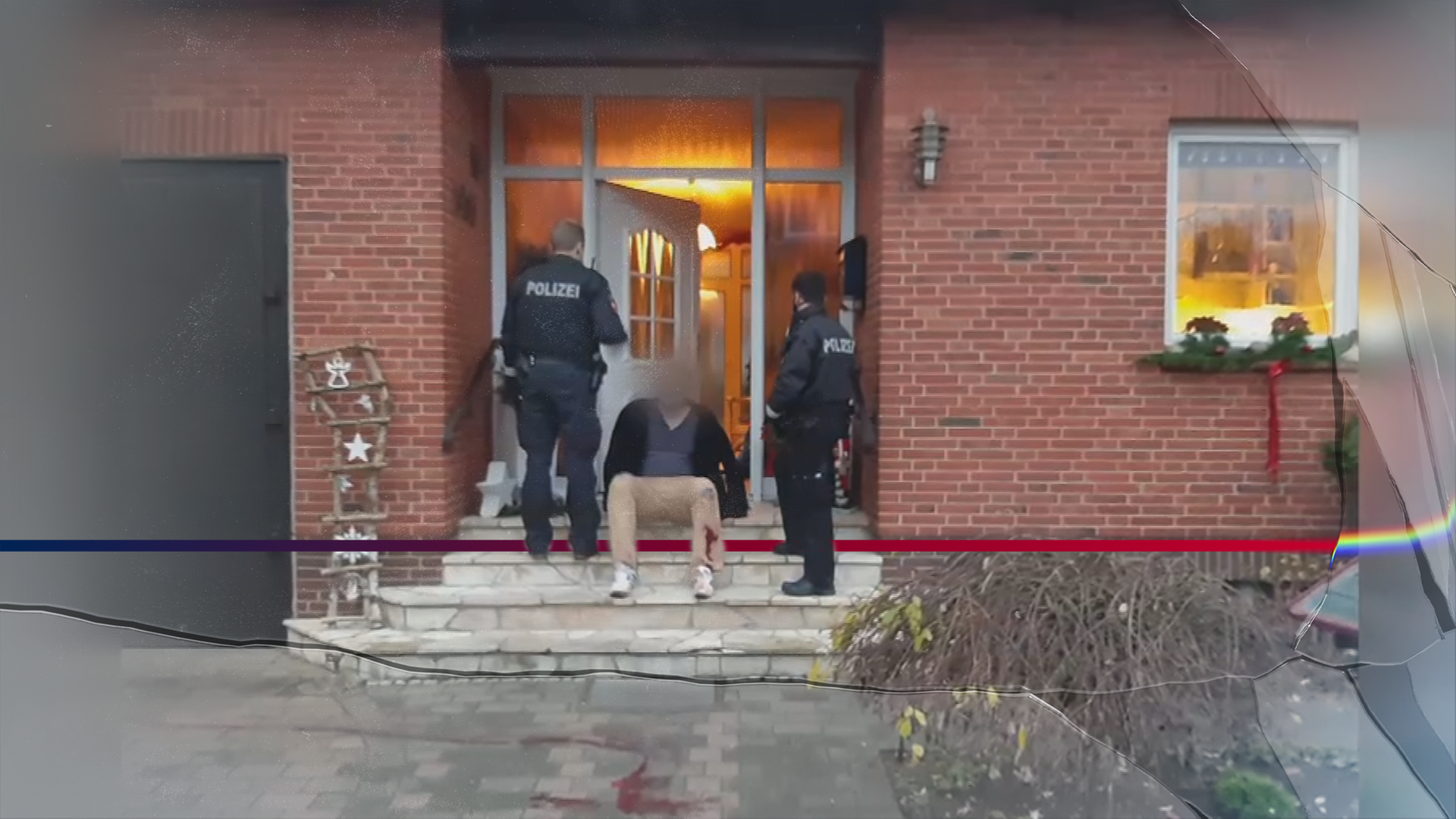 Ein Mann sitzt mit blutverschmierter Hose auf einer Hauseingangstreppe, bewacht von zwei Polizisten. Die Haustüre ist geöffnet. Auf der Treppe diverse Dekorationsartikel. Im Anschnitt noch zu sehen am rechten Bildrand: Ein Blumenbeet und ein Fenster. Die Beleuchtung im Haus ist an.