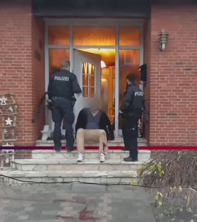 Ein Mann sitzt mit blutverschmierter Hose auf einer Hauseingangstreppe, bewacht von zwei Polizisten. Die Haustüre ist geöffnet. Auf der Treppe diverse Dekorationsartikel. Im Anschnitt noch zu sehen am rechten Bildrand: Ein Blumenbeet und ein Fenster. Die Beleuchtung im Haus ist an.