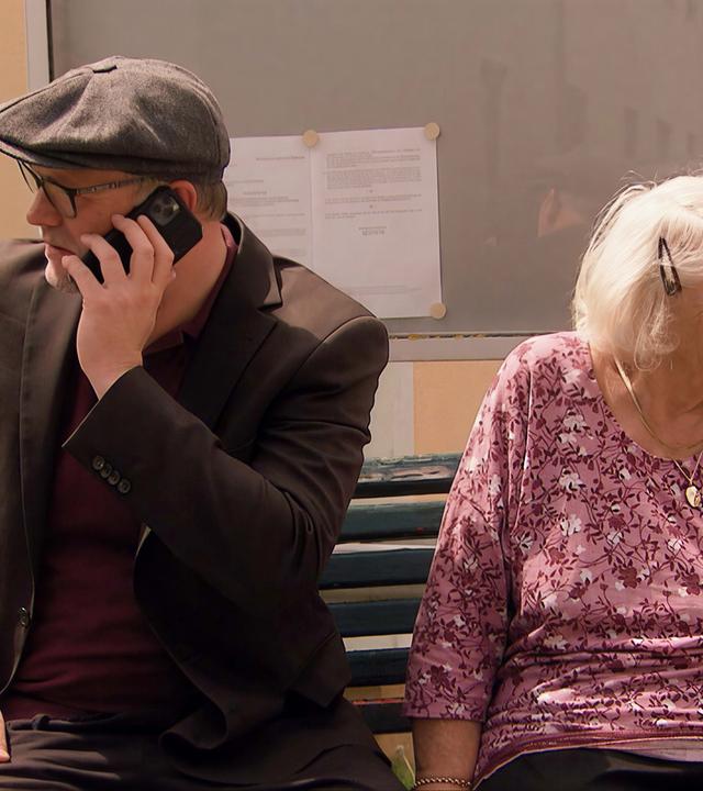 Das Bild zeigt eine Szene, die während der Dreharbeiten zu einer TV-Dokumentation stattfindet. Im Vordergrund sitzen zwei Personen auf einer Bank. Auf der linken Seite ist ein Mann zu sehen, der ein Handy in der Hand hält und telefoniert. Er trägt eine schwarze Jacke und einen grauen Hut. Auf der rechten Seite sitzt eine ältere Frau mit schneeweißem Haar, die ein rosa Shirt trägt und einen Gehstock dabei hat. Beide blicken in unterschiedliche Richtungen. Hinter ihnen ist eine Wand mit einem Fenster und einem Plakat oder einer Mitteilung, die an einem Brett befestigt ist. Die Umgebung vermittelt einen ländlichen Charakter, wahrscheinlich in einer idyllischen Gemeinde im Südburgenland.