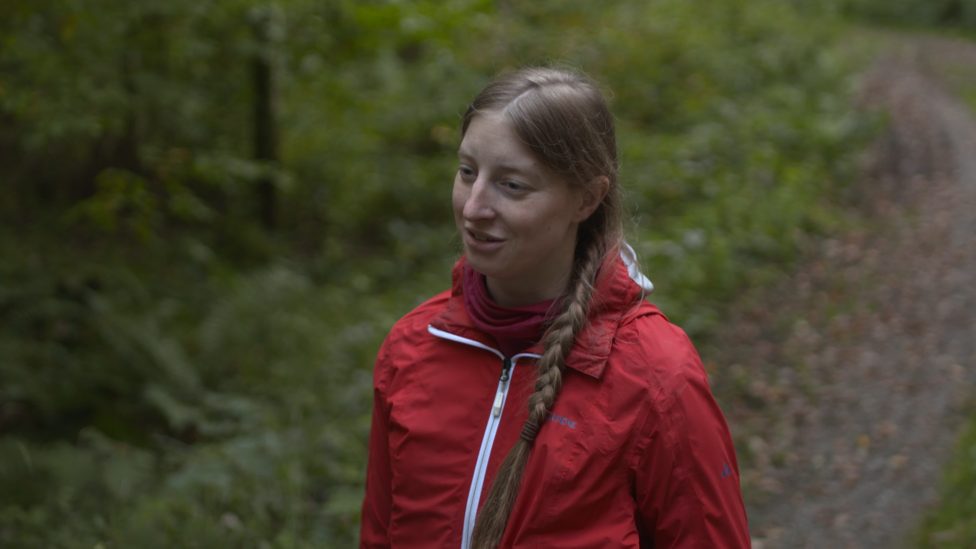 Eine junge, blonde Frau mit langem Flechtzopf steht in roter Outdoor-Jacke im Wald.