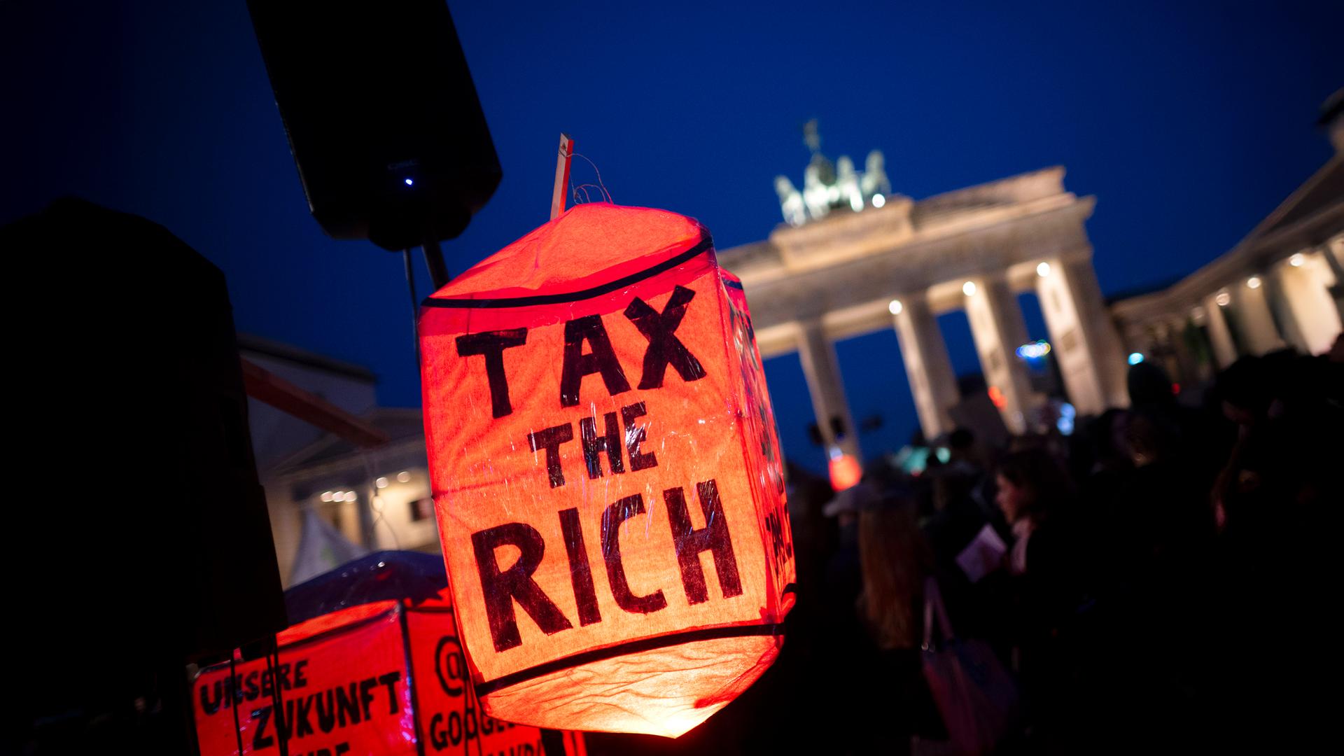 Bei einer Demo vor dem Brandenburger Tor bei Nacht ist im Vordergrund eine rote Lampe mit dem Text tax the rich zu sehen.