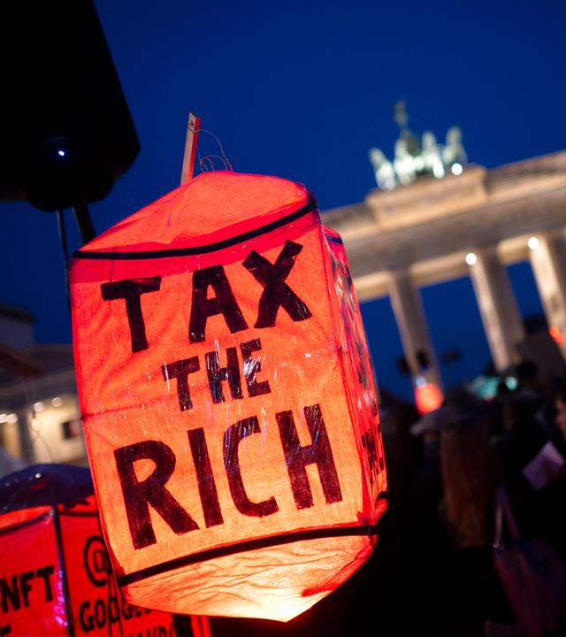 Bei einer Demo vor dem Brandenburger Tor bei Nacht ist im Vordergrund eine rote Lampe mit dem Text tax the rich zu sehen.