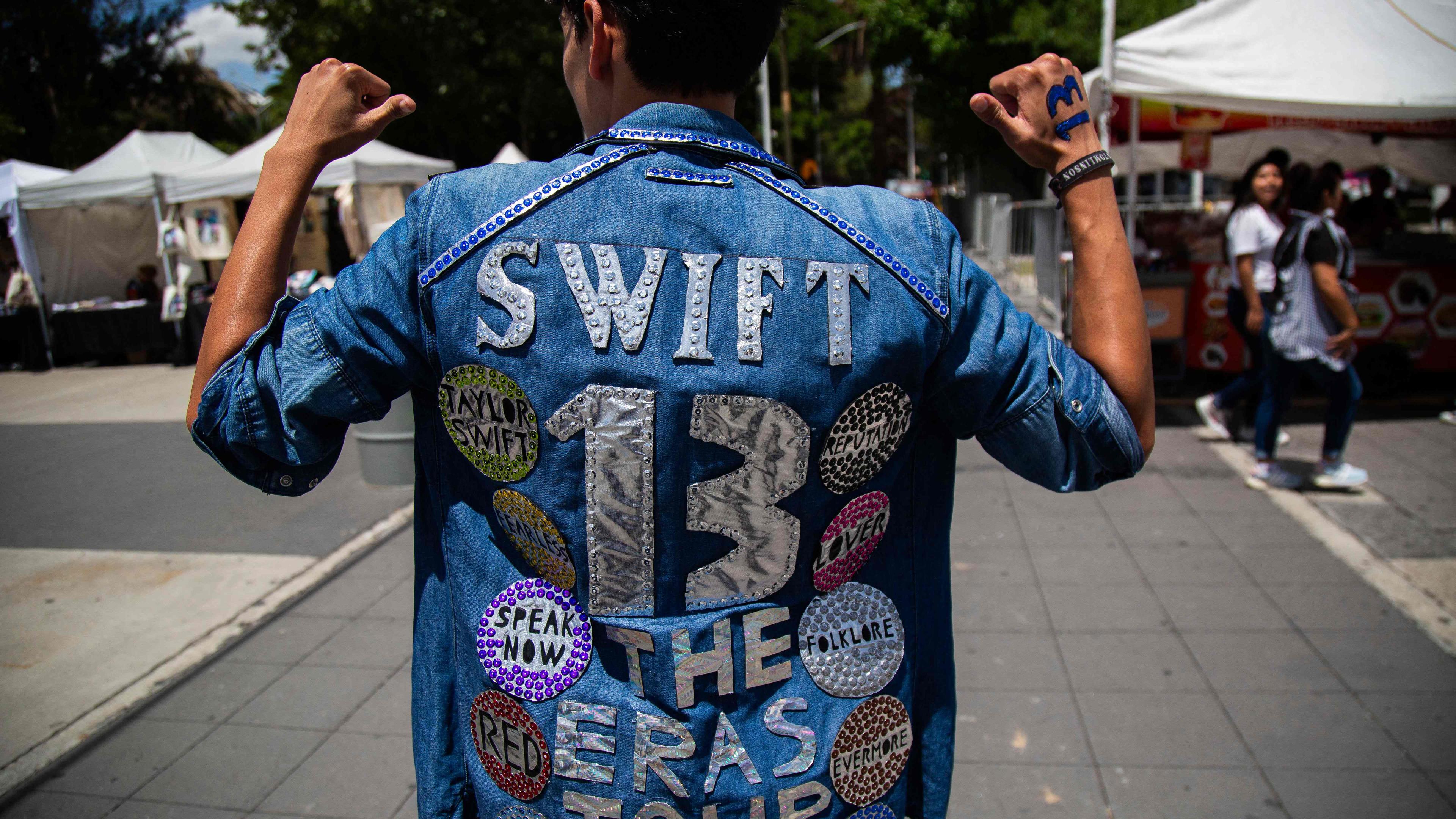 Eine Person, die auf die Rückseite ihrer Jeansjacke zeigt. Auf der Jacke sind Taylor-Swift-Patches.