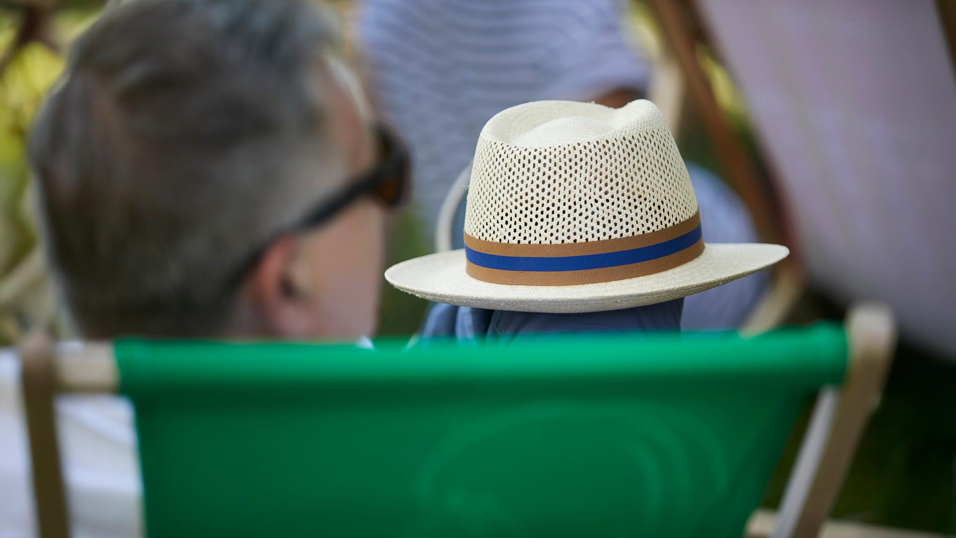Ein Mann mit grauen Haaren sitzt in einem grünen Liegestuhl und hält einen Strohhut mit blau-braunem Band auf dem Schoß; die Szene spielt sich im Garten ab.