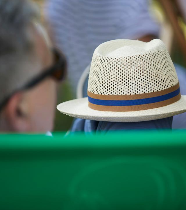 Ein Mann mit grauen Haaren sitzt in einem grünen Liegestuhl und hält einen Strohhut mit blau-braunem Band auf dem Schoß; die Szene spielt sich im Garten ab.