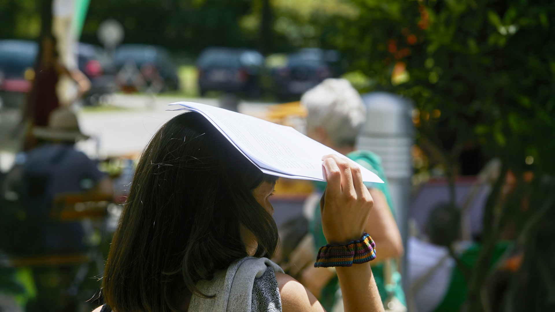 Eine Frau hält sich bei sommerlichem Wetter ein Blatt Papier über den Kopf, um sich vor der Sonne zu schützen; im Hintergrund sind weitere Personen im Garten zu sehen.