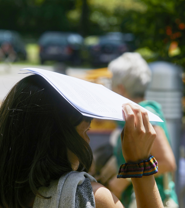 Eine Frau hält sich bei sommerlichem Wetter ein Blatt Papier über den Kopf, um sich vor der Sonne zu schützen; im Hintergrund sind weitere Personen im Garten zu sehen.