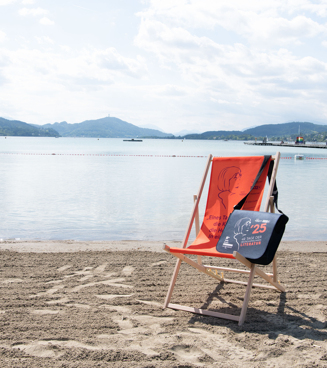 Liegestuhl und Tasche mit Bachmannpreis-Logo am Ufer eines Sees mit Bergen im Hintergrund