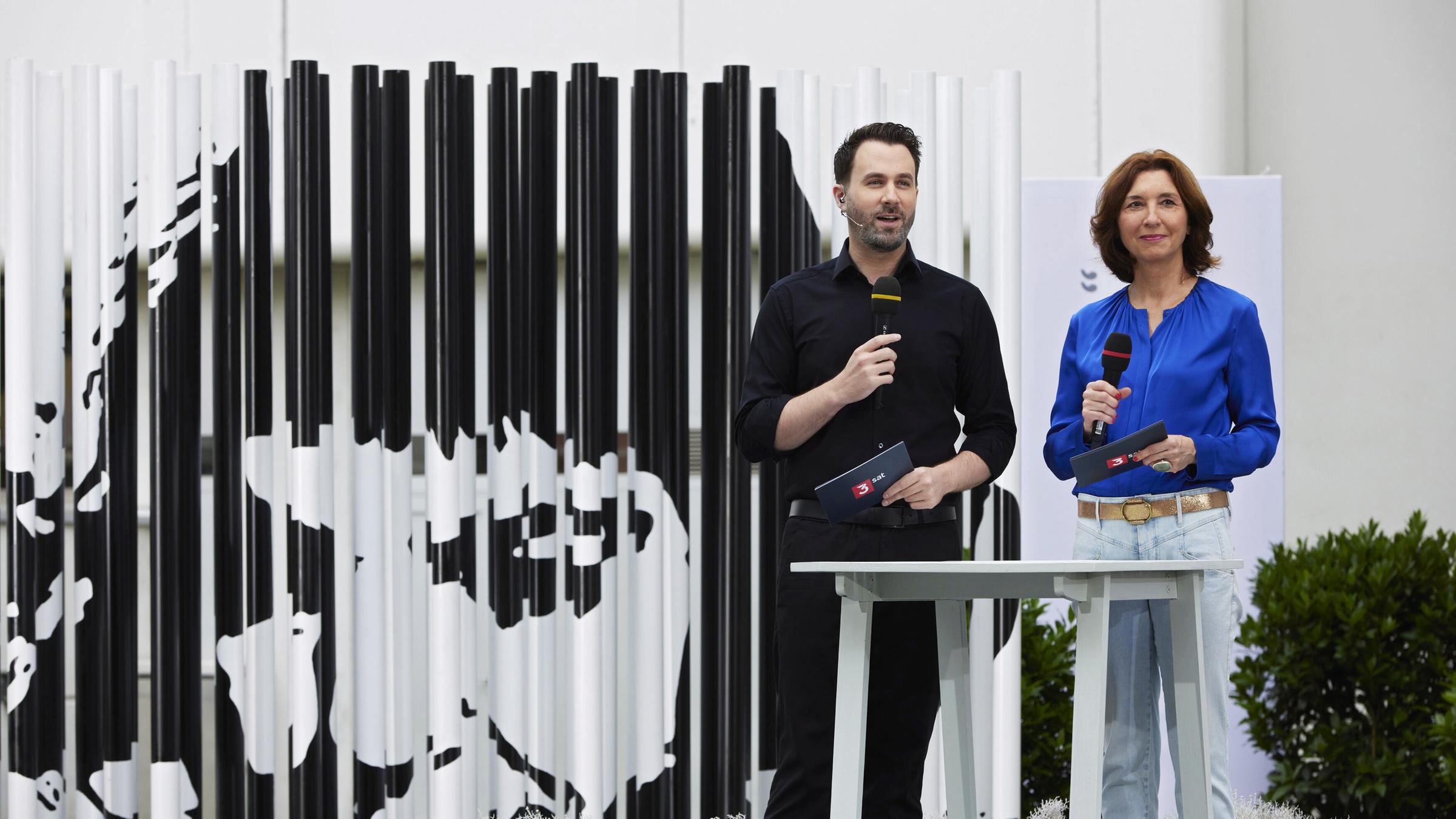 Ein Mann und eine Frau stehen mit Mikrofon vor einem abstrakten großen Portrait von Ingeborg Bachmann