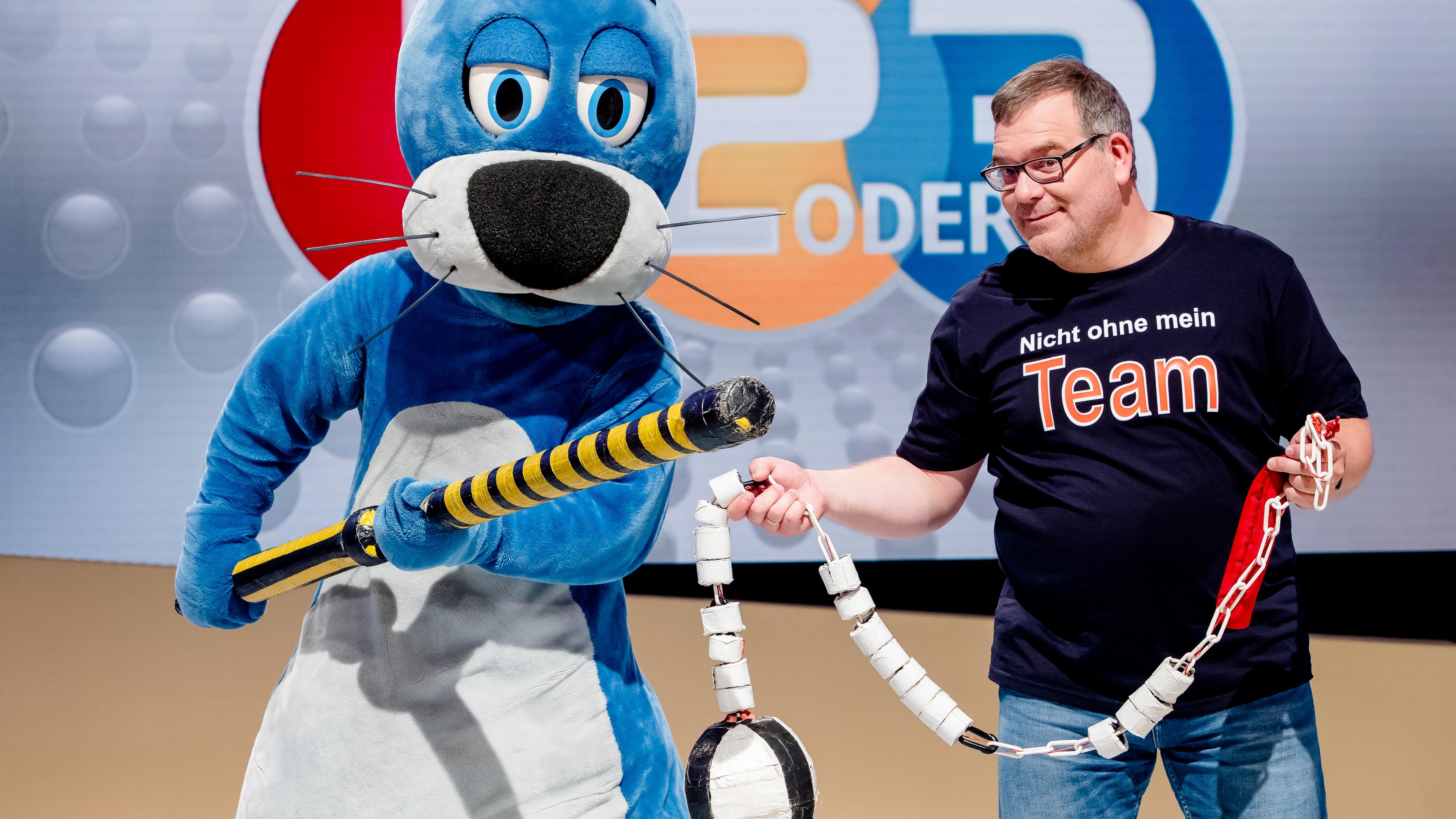 1, 2 oder 3: Moderator Elton steht mit Piet Flosse vor der Vidiwall im "1, 2 oder 3"-Studio. Beide halten Sportgeräte in der Hand.