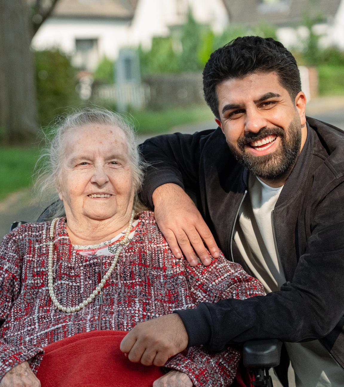 Junger Mann mit schwarzem Vollbart und schwarzen Haaren lehnt sich lachen auf den Rollstuhl einer sehr betagten Frau darin. Auch sie lacht in die Kamera.