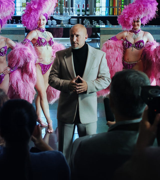 Gerd Kolberg (Benno Fürmann) steht vor einer Reihe Reportern und verkündet etwas. Hinter ihm posieren vier lächelnde Tänzerinnen in pinkfarbenen Kostümen vor einer Bar.