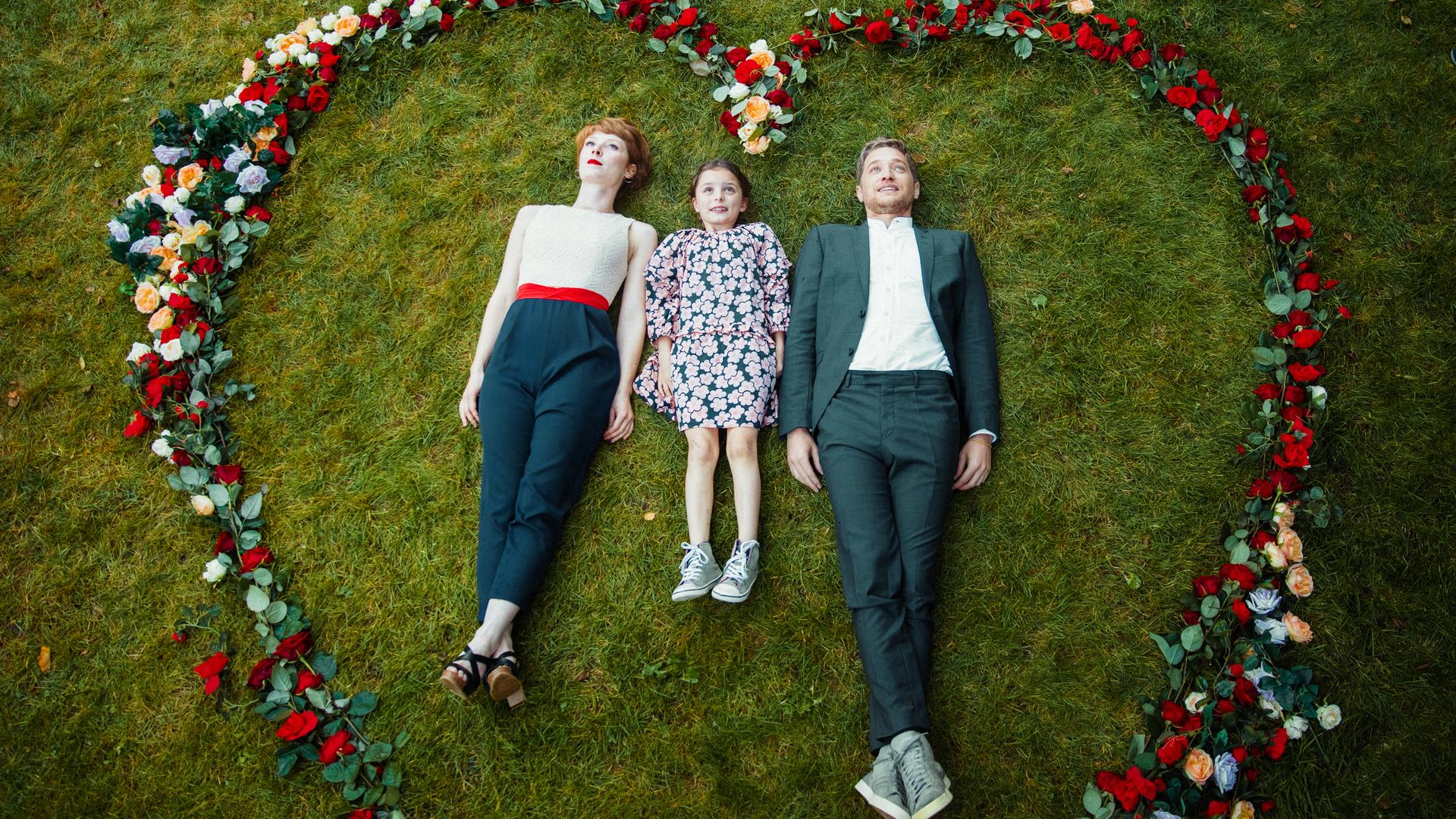 Lena (Sarah Bauerett), ihre Pflegetochter Talisa (Filippa Möller) und ihr Verlobter Marek (Markus “Maeckes” Winter) machen das Foto für ihre Hochzeitseinladungskarte. Dafür liegen sie auf dem Rasen, in einem Herz aus Rosen.