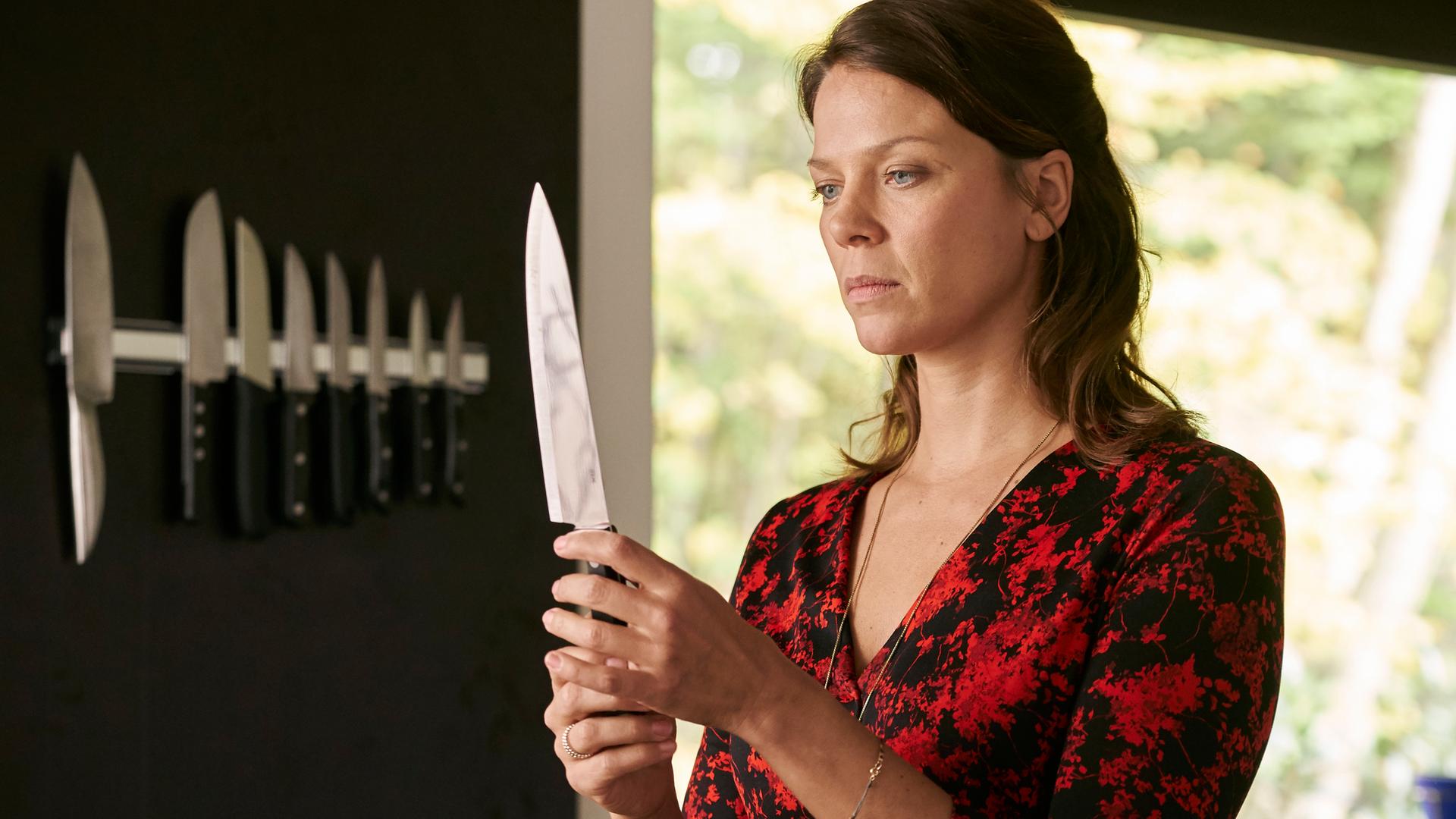  "Hattinger und der Nebel": Sarah Beck (Jessica Schwarz) steht in einem Raum und hält mit beiden Händen ein sehr spitzes Messer, auf das sie nachdenklich starrt. An der Wand hängt eine Magnet-Leiste mit weiteren Küchenmessern.