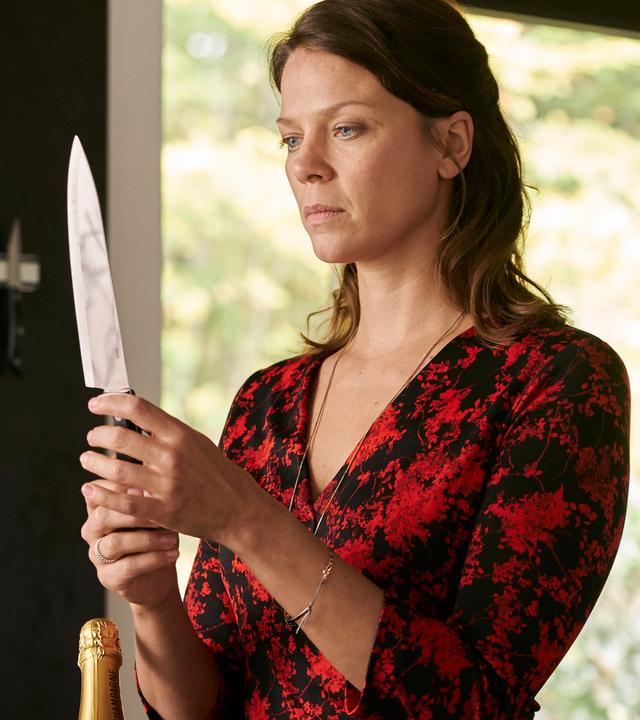  "Hattinger und der Nebel": Sarah Beck (Jessica Schwarz) steht in einem Raum und hält mit beiden Händen ein sehr spitzes Messer, auf das sie nachdenklich starrt. An der Wand hängt eine Magnet-Leiste mit weiteren Küchenmessern.