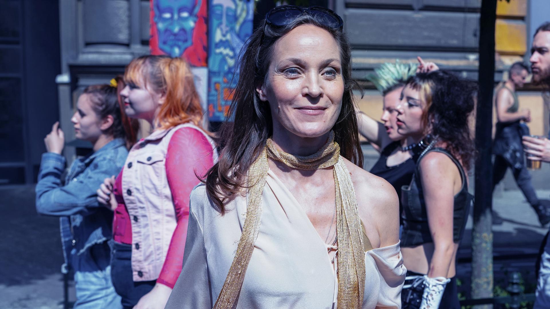 Regina Feldmann (Jeanette Hain) steht auf einer Straße umringt von Besuchern der Loveparade. Sie lächelt jemanden außerhalb des Bildes zu.