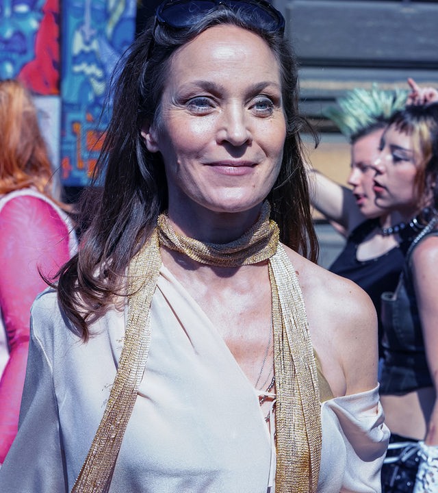 Regina Feldmann (Jeanette Hain) steht auf einer Straße umringt von Besuchern der Loveparade. Sie lächelt jemanden außerhalb des Bildes zu.