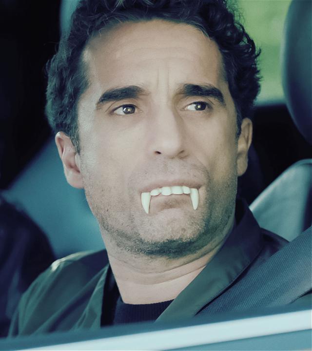 Ina Behrendsen (l., Julia Brendler) und Hinnerk Feldmann (r., Oliver Wnuk) sitzen in einem Auto und schauen durch das geöffnete Fenster auf der Fahrerseit. Hinnerk trägt ein Vampirgebiss aus Plastik. Er hat zwar den Mund geschlossen, doch die Vorderzähne ragen zwischen seinen Lippen hervor.