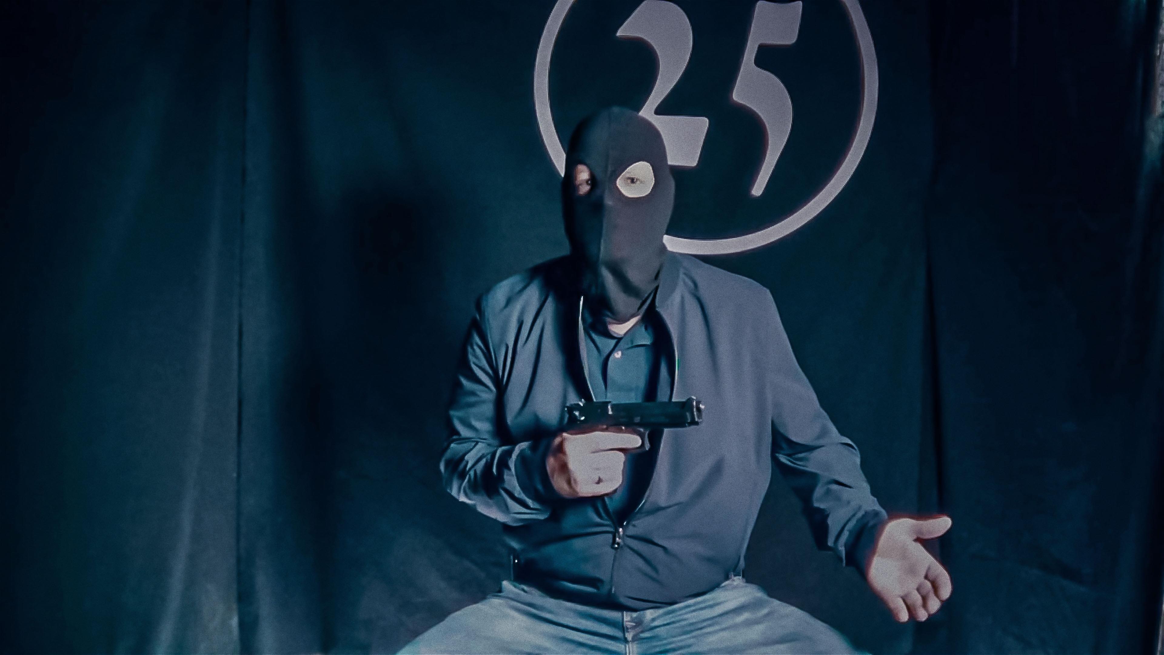 Ein Mann in einer Sturmmaske und mit einer Pistole in der Hand sitzt vor einem schwarzen Vorhang, auf dem die Zahl 25 aufgenäht ist. Das Bild sieht etwas verwaschen aus, als ob es aus einem Onlinevideo oder Livestream stammt.