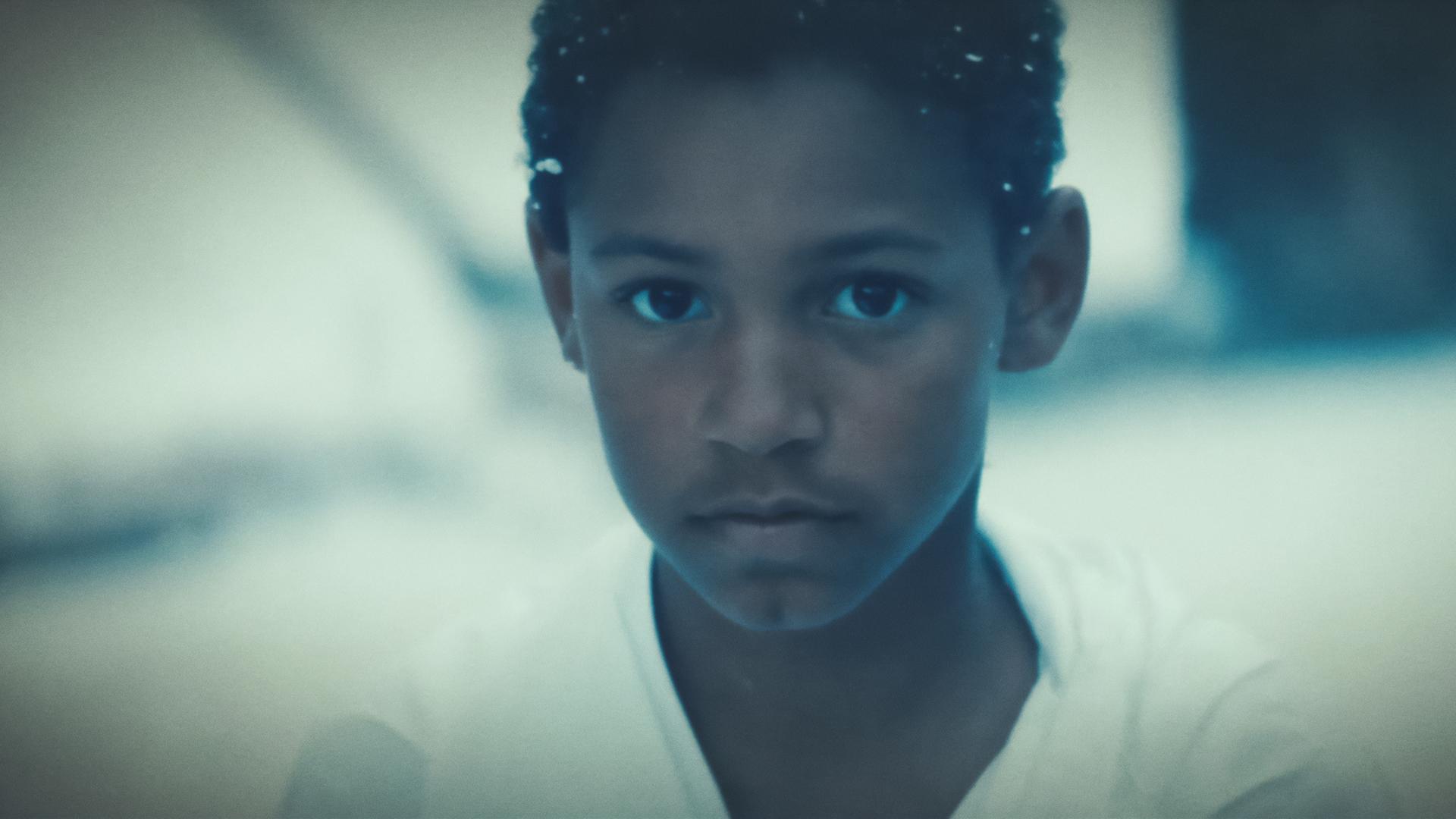 Jonny (Joel A.), ein circa 10-jähriger schwarzer Junge, steht in einem weißen T-Shirt im Schnee. 