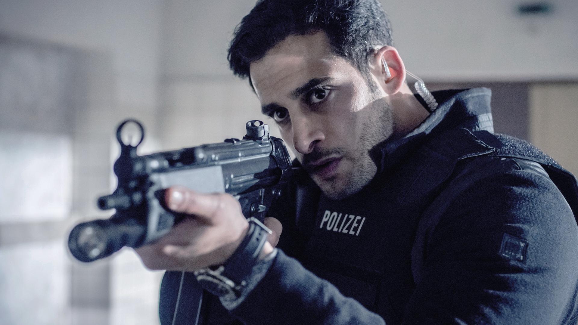 Karim Uthman (Karim Günes) trägt eine schusssichere Polizeiweste und zielt mit einem Gewehr auf etwas außerhalb des Bildes.