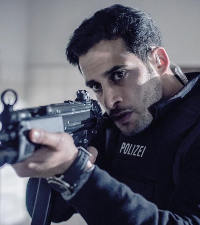 Karim Uthman (Karim Günes) trägt eine schusssichere Polizeiweste und zielt mit einem Gewehr auf etwas außerhalb des Bildes.