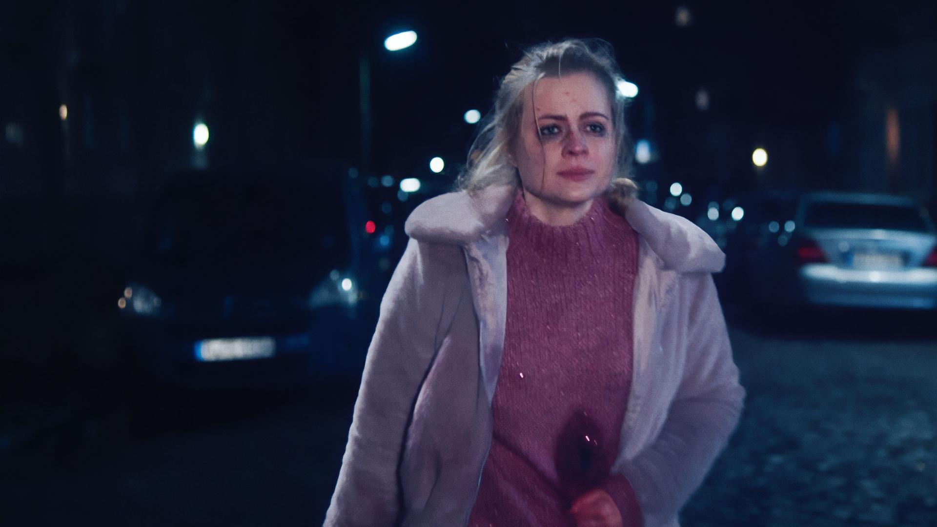 Eine junge Frau (Kara Edenberg) läuft eine nächstliche Straße hinunter. Sie hat Blutergüsse im Gesicht und hält sich den Bauch, wo man eine blutende Wunde sehen kann.