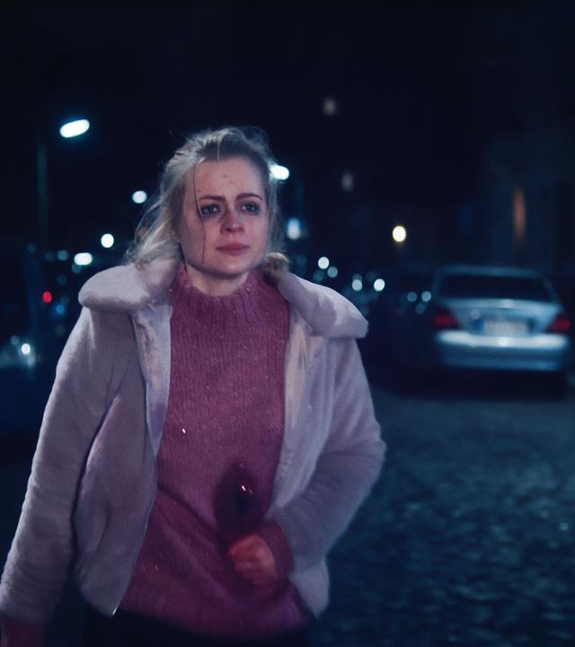 Eine junge Frau (Kara Edenberg) läuft eine nächstliche Straße hinunter. Sie hat Blutergüsse im Gesicht und hält sich den Bauch, wo man eine blutende Wunde sehen kann.