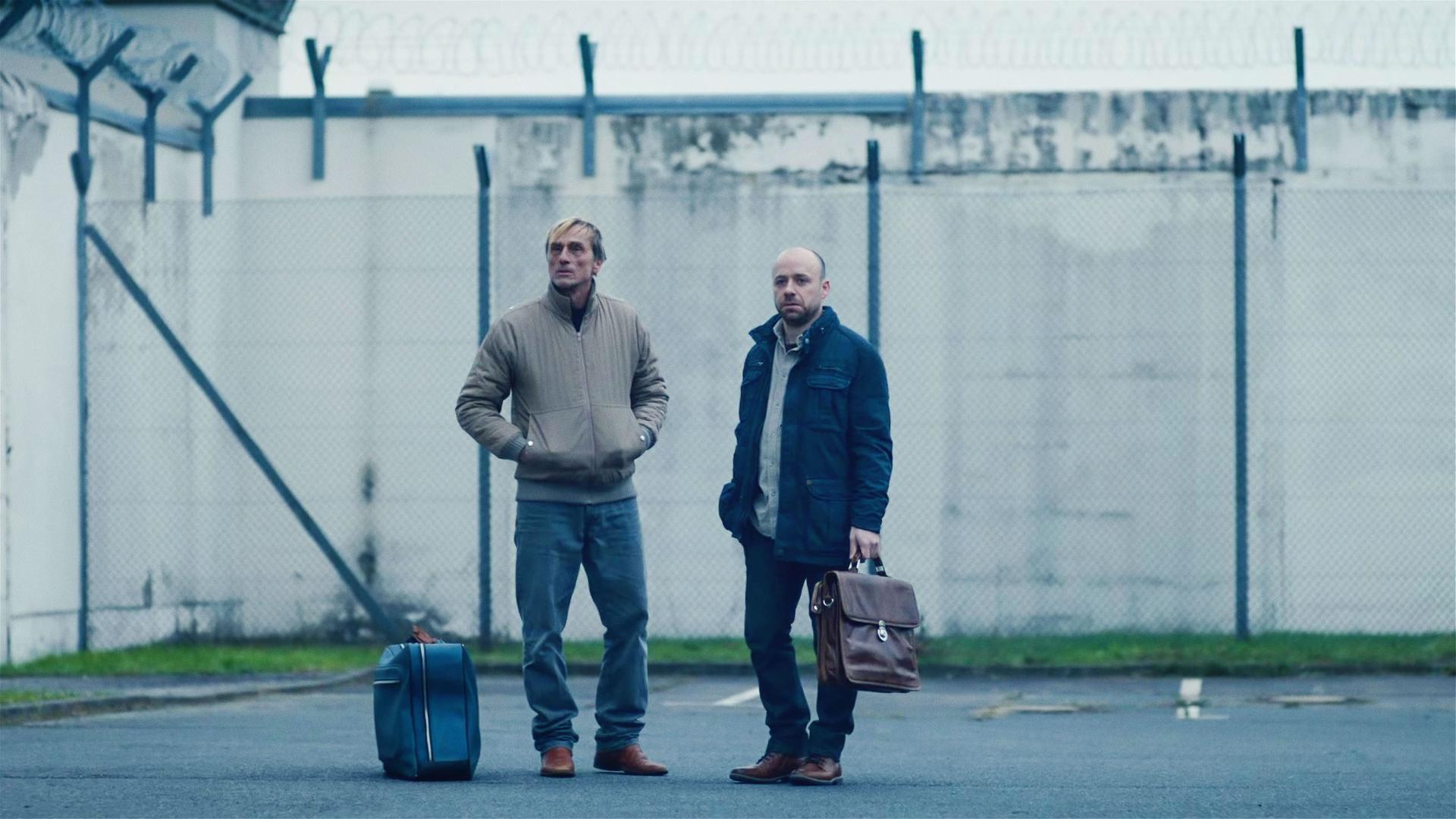 Zwei Männer stehen mit Koffern in der Hand im Innenhof eines Gefängnisses.