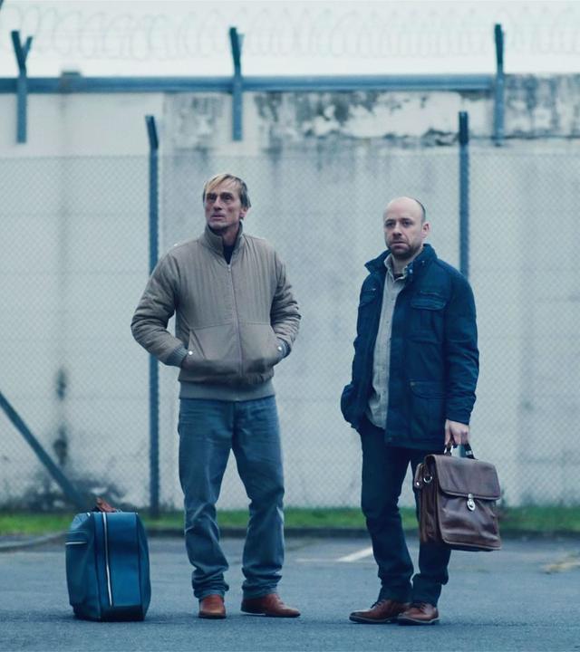 Zwei Männer stehen mit Koffern in der Hand im Innenhof eines Gefängnisses.