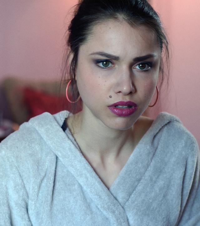 Jemma Dubnitzki (Vivien Sczesny) in Nahaufnahme. Jemma sitzt im Bademantel auf ihrem Bett und schaut bestürzt auf das Geschehen. Auf ihrem Bett steht ein Schild der Spurensicherung.