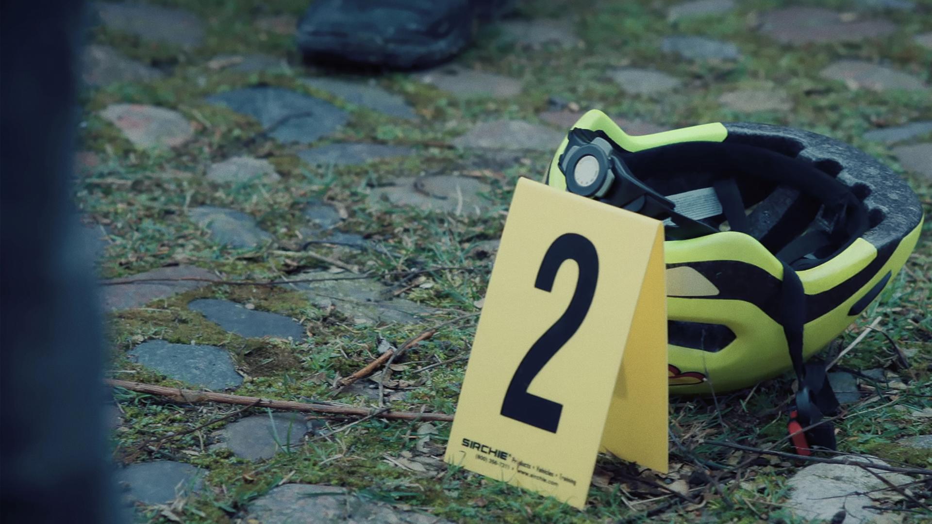 Auf einem mit grasbewachsenen Weg aus Kopfsteinplaster liegt ein neongelber Fahrradhelm. Daneben steht ein Aufsteller zum Kennzeichnen von Beweismitteln mit der Nummer 2 darauf.